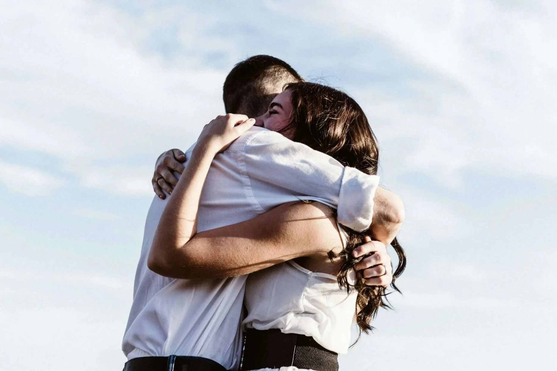 A man and woman hugging each other outdoors against a cloudy sky.