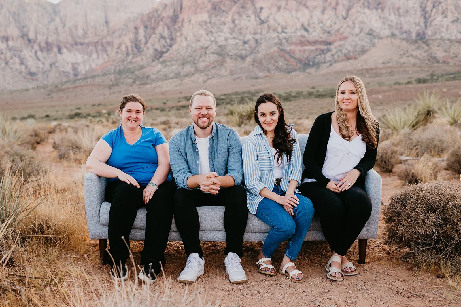Four people sitting on a grey sofa outdoors in a desert landscape with mountains and sparse vegetation in the background.