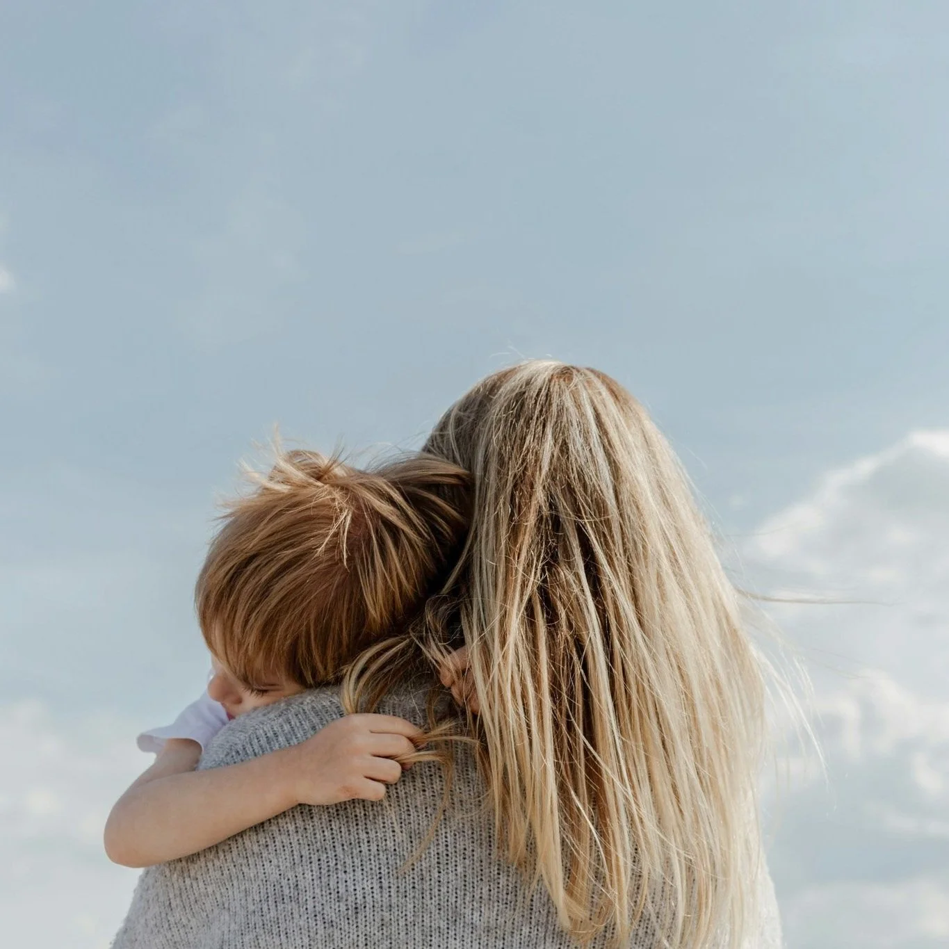 A woman hugging a young child outdoors with a partly cloudy sky in the background.