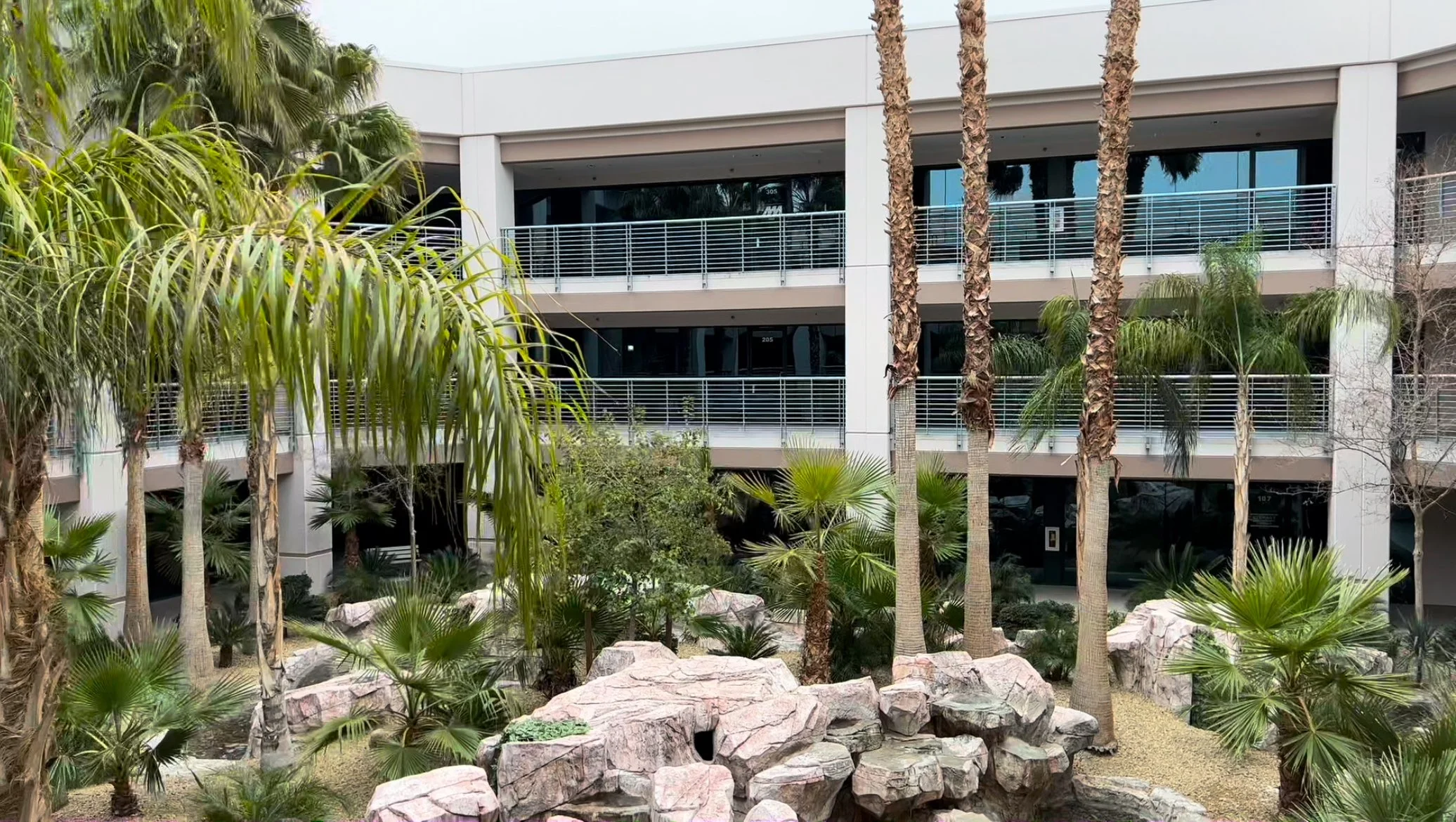 Modern office building with glass windows and balconies, surrounded by palm trees and a landscaped garden with rocks and greenery.