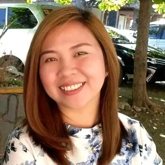 A young woman with shoulder-length reddish-brown hair smiling outdoors with trees and parked cars in the background.