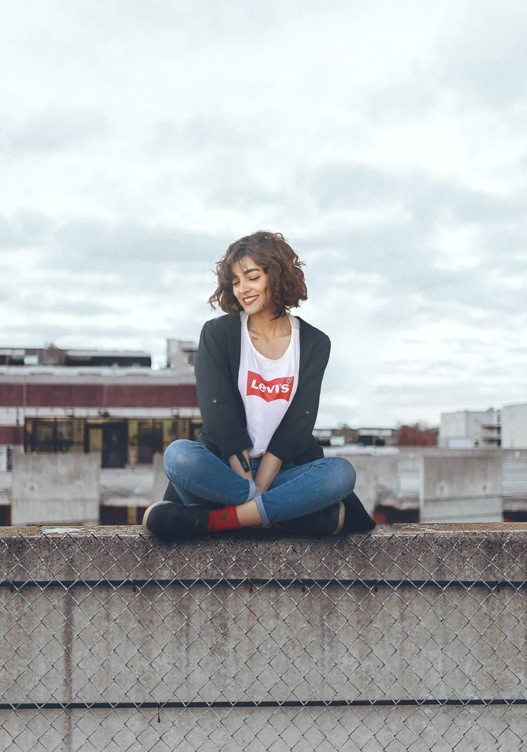 A young woman with curly brown hair sits cross-legged on a rooftop wall with a cityscape and cloudy sky in the background.